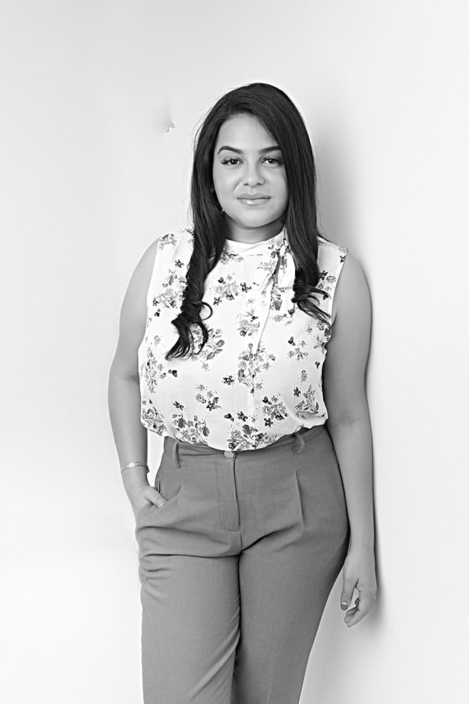 A woman with long dark hair stands against a plain background, wearing a sleeveless floral blouse and high-waisted trousers, with one hand in her pocket and a neutral expression. The photo is in black and white. - Jodi O Photography