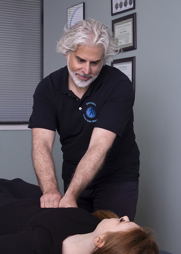 A man with gray hair and a beard, wearing a black polo shirt, performs a chiropractic adjustment on a woman lying on a table in a medical office with framed certificates on the wall. - Jodi O Photography