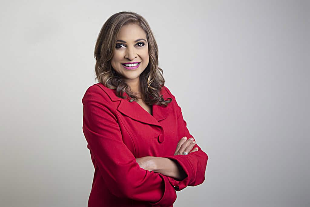 A woman with long brown hair smiles confidently, standing with arms crossed. She is wearing a bright red blazer and is posed against a plain, light gray background. - Jodi O Photography