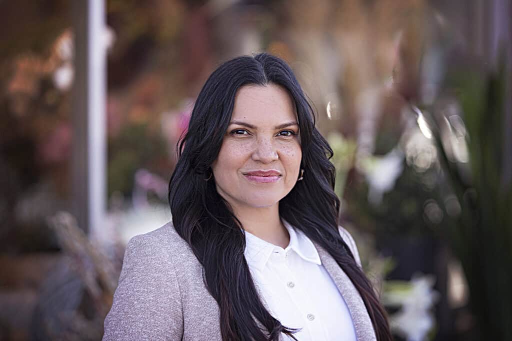 A woman with long dark hair, wearing a white blouse and light-colored blazer, stands outdoors smiling softly. The background is blurred with greenery and warm tones. - Jodi O Photography