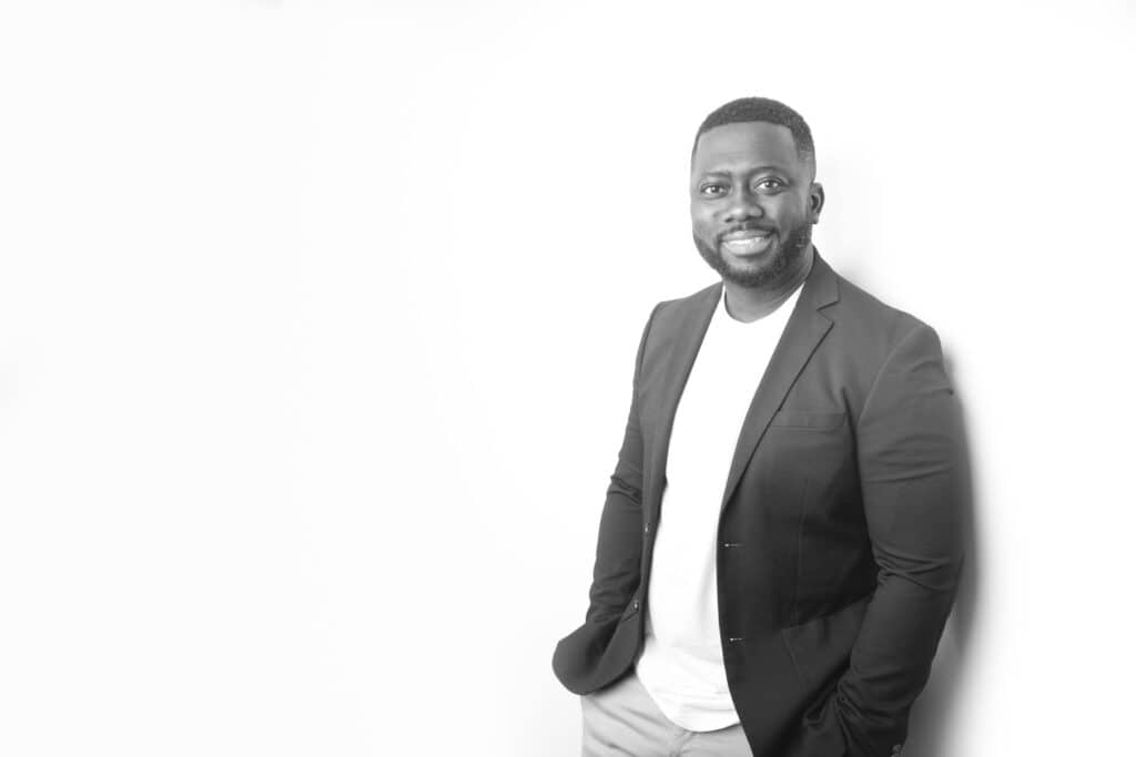 A man in a blazer and T-shirt smiles while leaning against a white wall, hands in his pockets. This black-and-white image with a bright, minimal background is perfect for men’s professional headshots or Calgary headshots. - Jodi O Photography