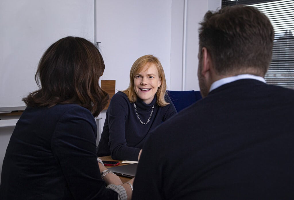 Three people sit around a table in an office, engaged in conversation. A blonde woman in a black sweater is smiling at two colleagues, who are seated with their backs to the camera. The atmosphere appears friendly and professional. - Jodi O Photography