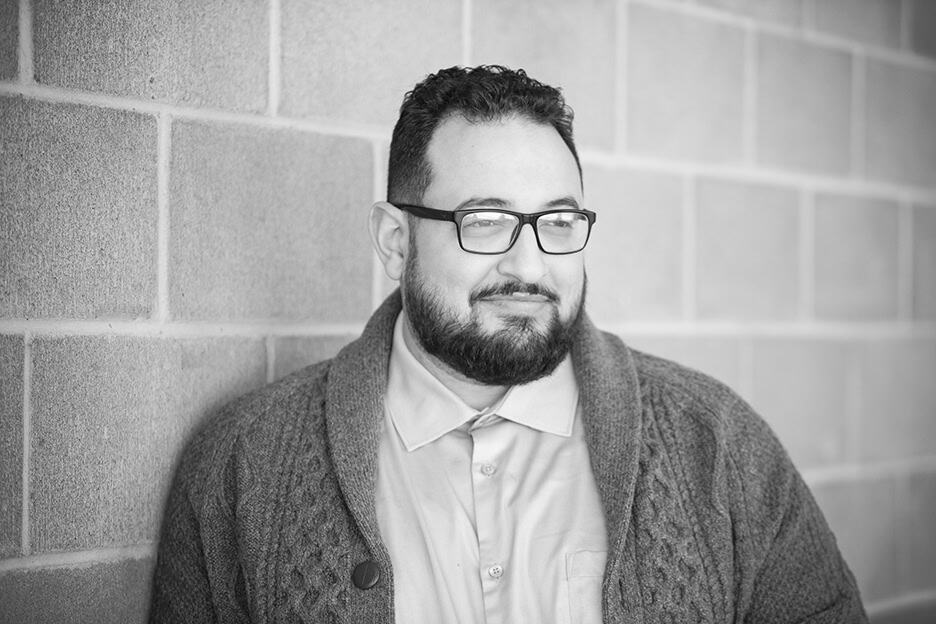 A man with dark hair, glasses, and a beard stands against a brick wall, wearing a collared shirt and a textured cardigan, looking slightly to the side and smiling gently. The image is in black and white. - Jodi O Photography