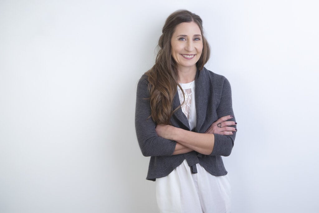 A woman with long brown hair, wearing a grey cardigan over a white blouse, stands against a plain white background with her arms crossed and smiling at the camera. - Jodi O Photography