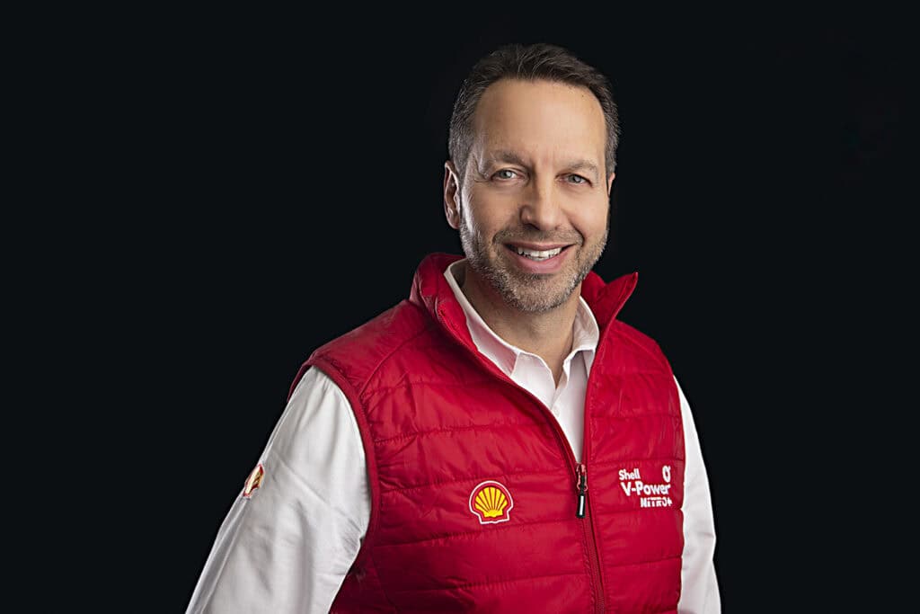 A man wearing a white shirt and a red Shell V-Power branded vest smiles in front of a black background. - Jodi O Photography