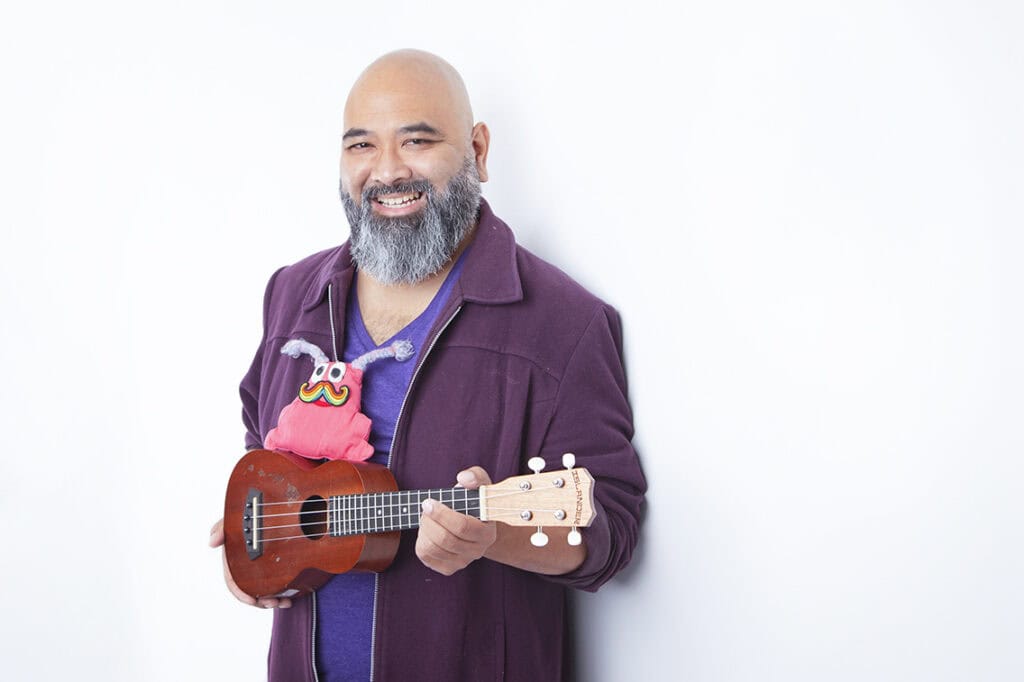 A smiling man with a gray beard, wearing a purple jacket, holds a ukulele with a colorful, playful puppet attached to it, standing against a plain white background. - Jodi O Photography