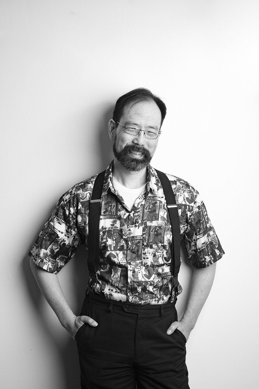 A man with glasses and a beard stands against a plain wall, wearing a patterned shirt, suspenders, and dark pants. He has his hands in his pockets and a slight smile on his face. - Jodi O Photography