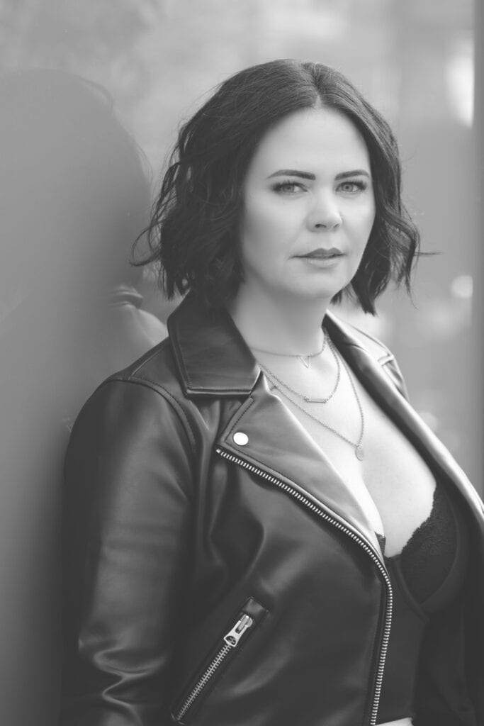 A woman with shoulder-length dark hair wearing a leather jacket and layered necklaces looks at the camera. The image is in black and white, with a blurred background. - Jodi O Photography