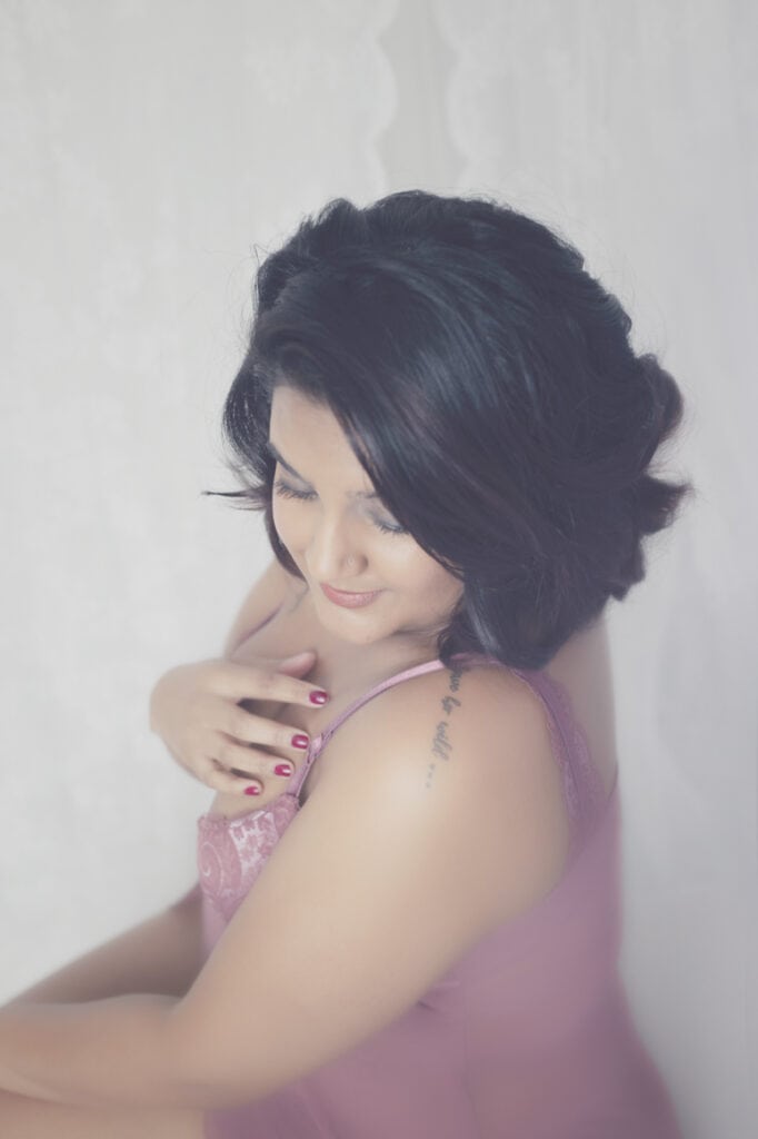 A woman with short, dark hair wearing a pink lace top looks down and smiles softly, with one hand touching her shoulder. The background is soft and white, giving the image an ethereal feel. - Jodi O Photography