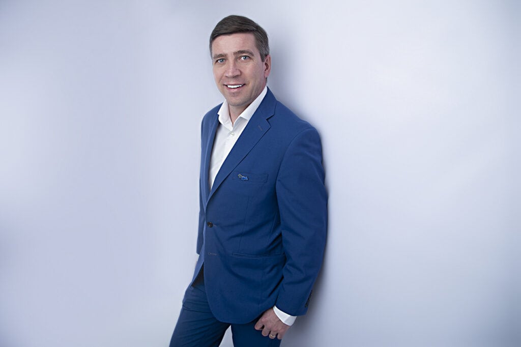A man in a blue suit and white shirt stands, smiling, against a plain light background. - Jodi O Photography