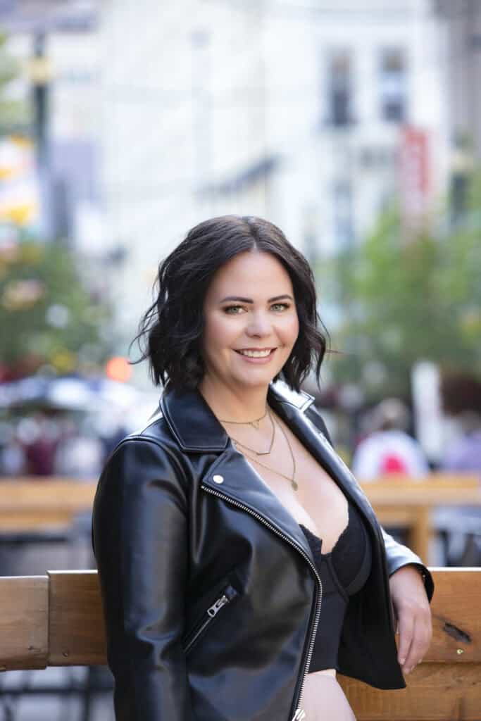 A woman with shoulder-length dark hair, wearing a black leather jacket and layered necklaces, smiles while leaning on a wooden railing in an outdoor urban setting with blurred buildings and trees in the background. - Jodi O Photography