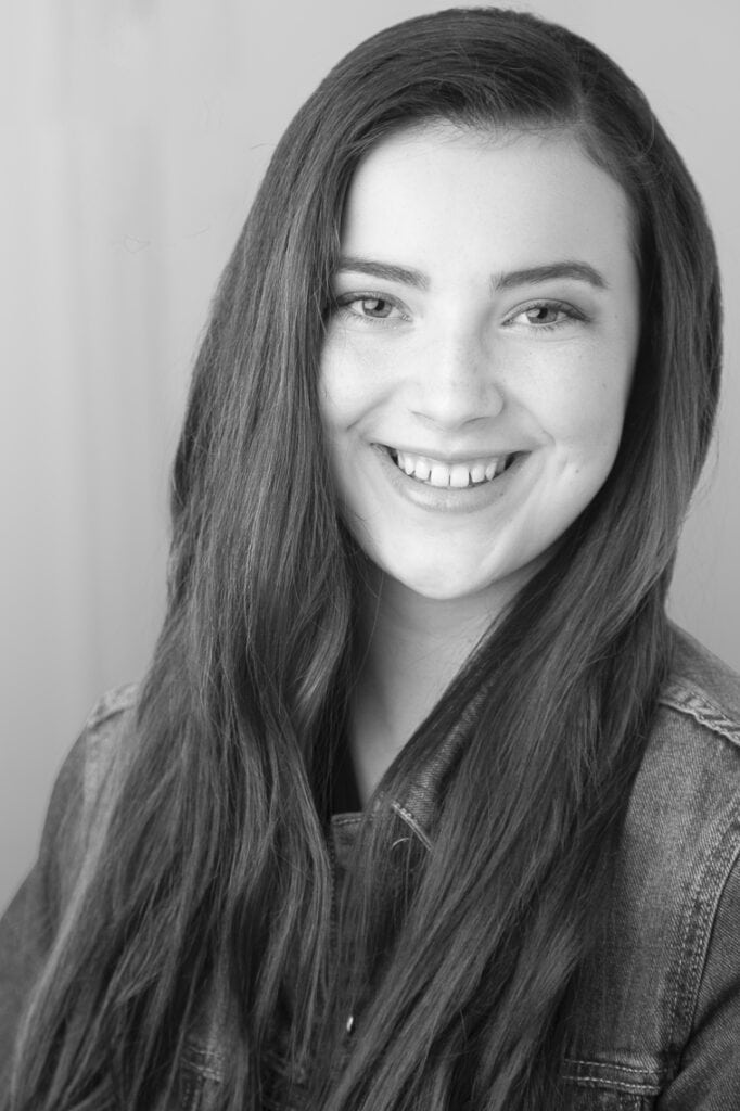 A young woman with long, straight hair smiles at the camera. She is wearing a denim jacket and the photo is in black and white, with a softly blurred background. - Jodi O Photography