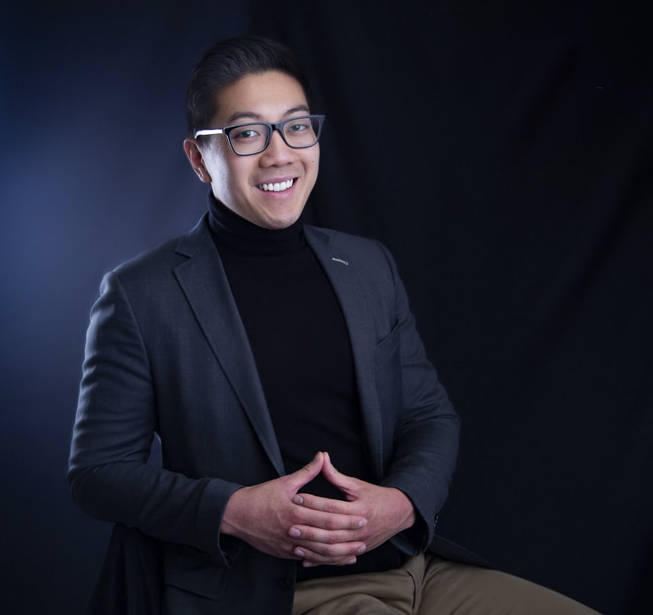 A man in glasses and a dark blazer sits and smiles in front of a black background. He wears a black turtleneck and tan pants, with his hands loosely clasped in front of him. - Jodi O Photography