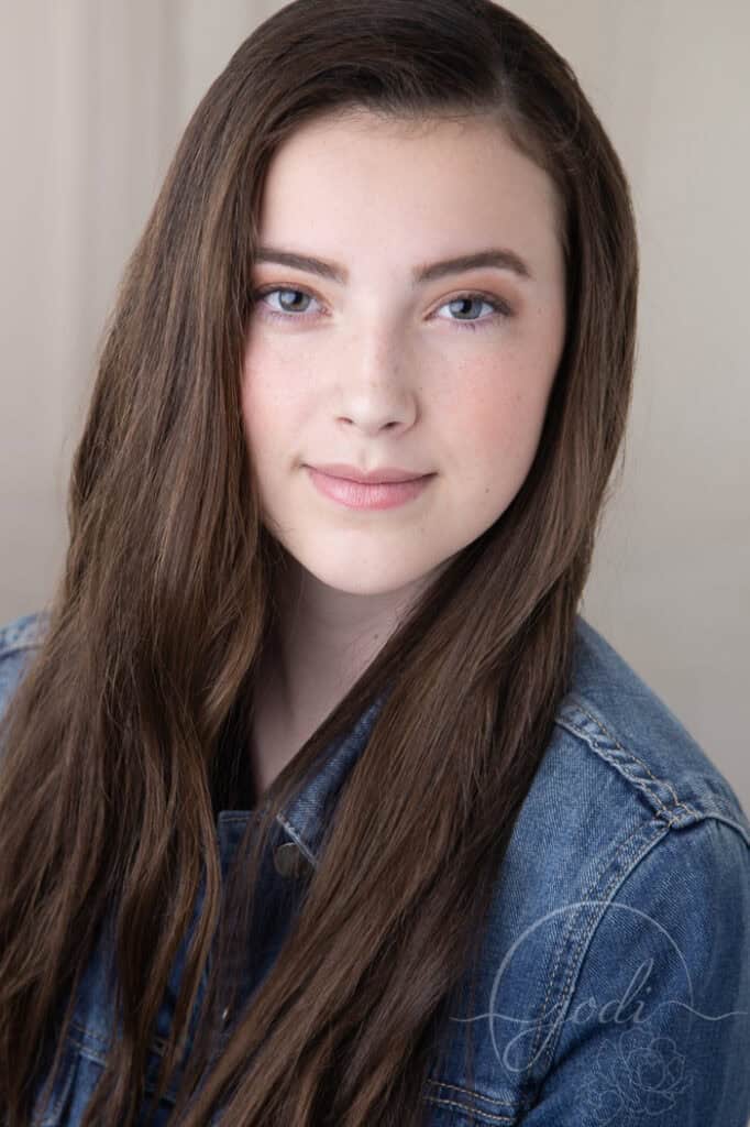 Teen girl with long brown hair and light skin, wearing a denim jacket, smiling softly and looking directly at the camera against a neutral background. - Jodi O Photography