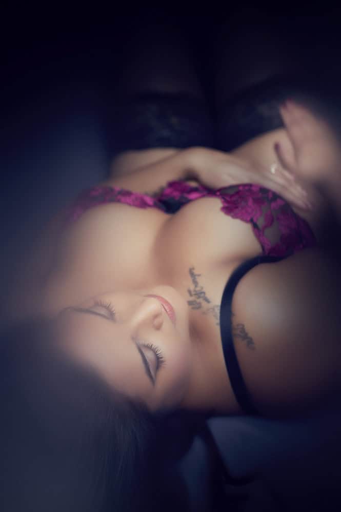 A woman wearing pink lace lingerie and black stockings lies on her back with her eyes closed, hands resting on her body. The image has a soft, dreamy focus and shows a tattoo on her chest. - Jodi O Photography