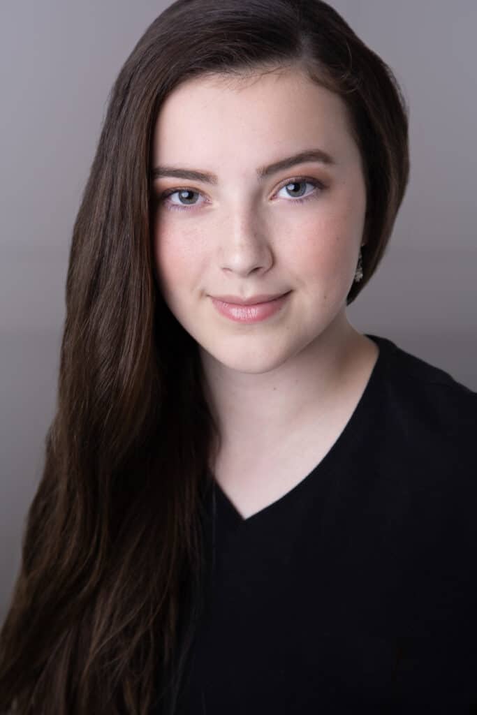 A young woman with long brown hair and blue eyes, wearing a black V-neck top, smiles softly at the camera against a plain, light background. - Jodi O Photography