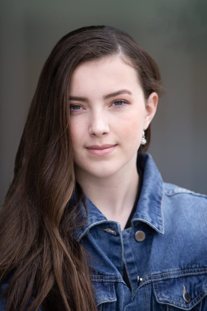 A young woman with long brown hair wearing a denim jacket looks directly at the camera with a neutral expression. The background is blurred. - Jodi O Photography
