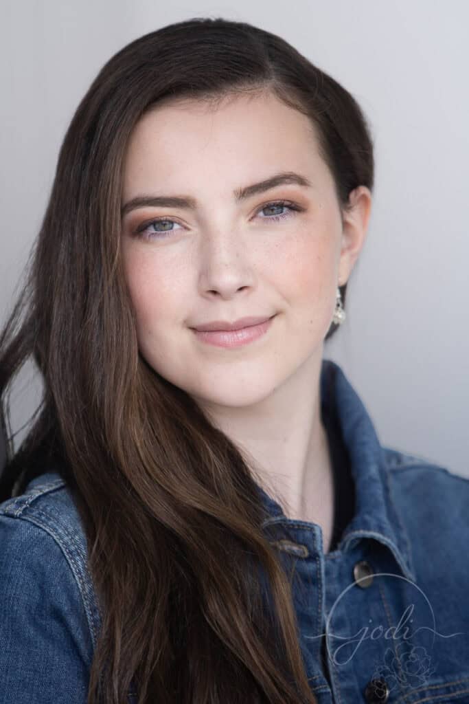 A young woman with long brown hair and light makeup smiles softly at the camera. She is wearing a denim jacket and small earrings, with a neutral background behind her. - Jodi O Photography