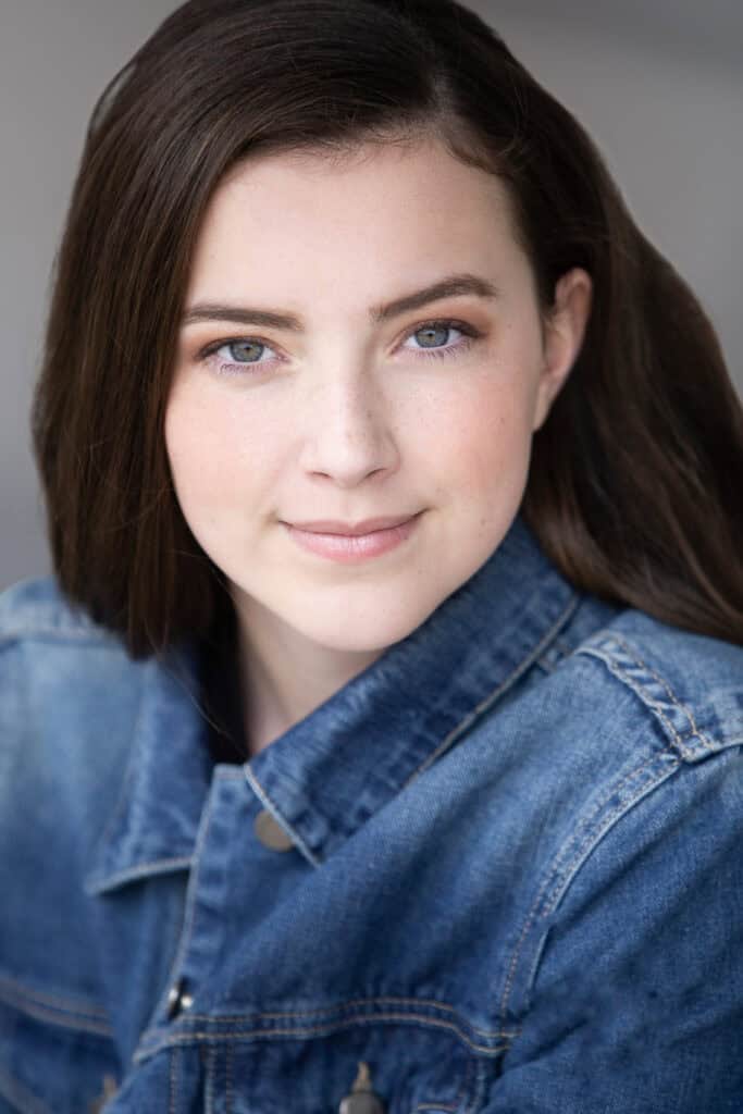 A young woman with fair skin, straight brown hair, and blue eyes smiles softly at the camera. She is wearing a blue denim jacket and is set against a neutral, blurred background. - Jodi O Photography