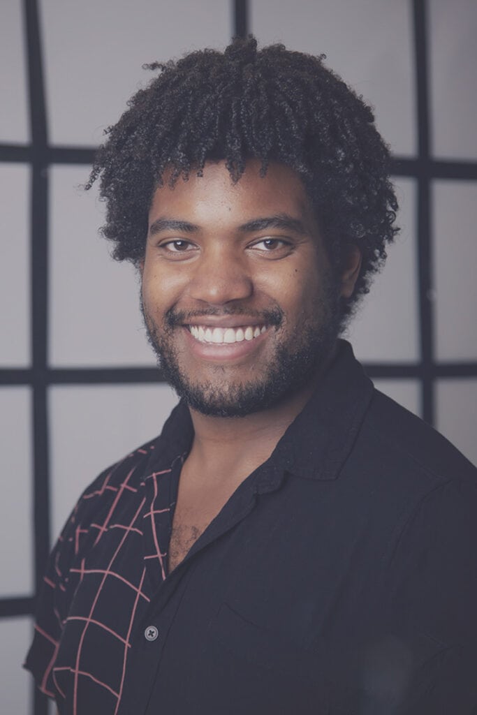 A smiling person with short curly hair and a beard, wearing a black shirt with a white and red grid pattern, stands in front of a light gray background with a bold black grid design. - Jodi O Photography