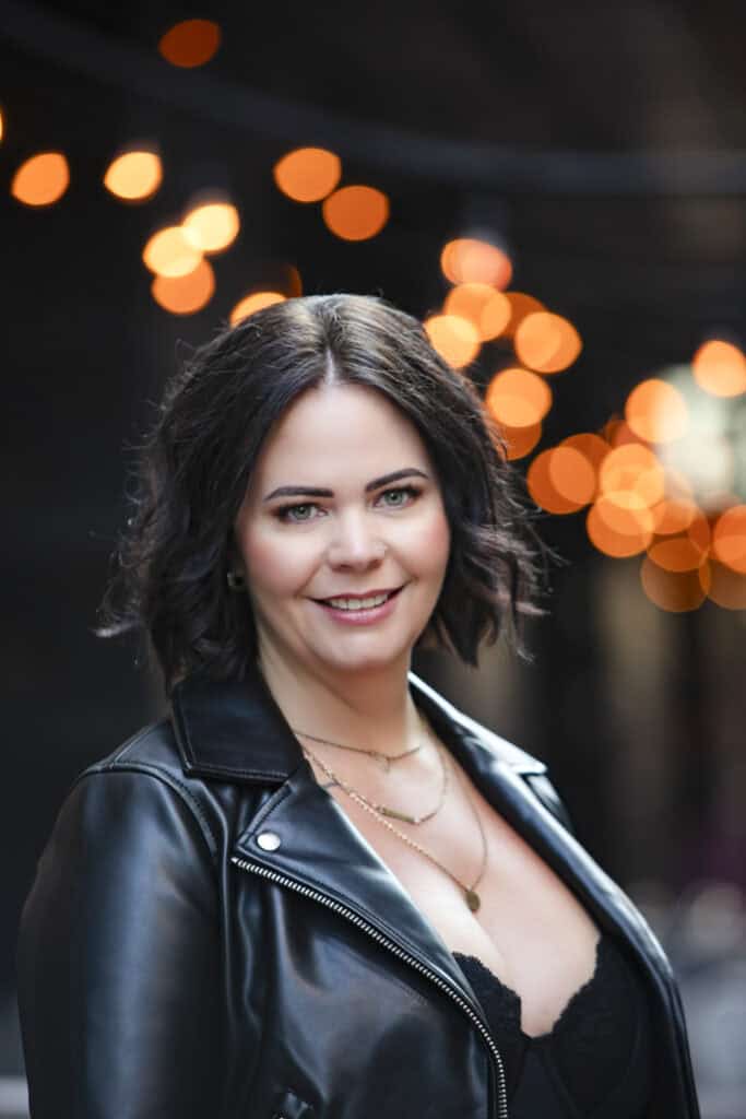 A woman with wavy dark hair and light eyes smiles at the camera, wearing a black leather jacket and gold necklaces. Warm bokeh lights glow softly in the blurred background. - Jodi O Photography