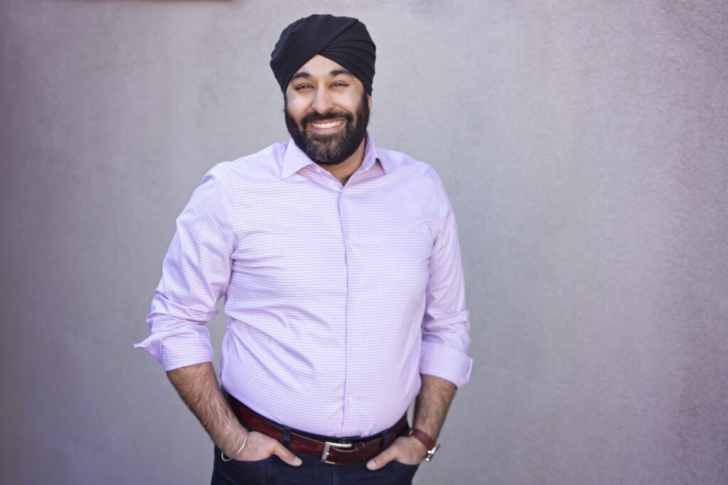 A man wearing a black turban and light pink button-up shirt smiles at the camera. He stands against a plain light-colored wall with his hands in his pockets. - Jodi O Photography