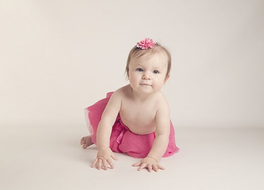 A baby wearing a pink flower headband and a pink skirt crawls on a light-colored floor, looking forward with a gentle expression. The background is plain and neutral. - Jodi O Photography