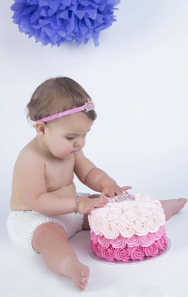 A baby wearing a pink headband and white shorts sits on the floor, touching a pink and white rosette cake, with a large purple decoration hanging in the background. - Jodi O Photography