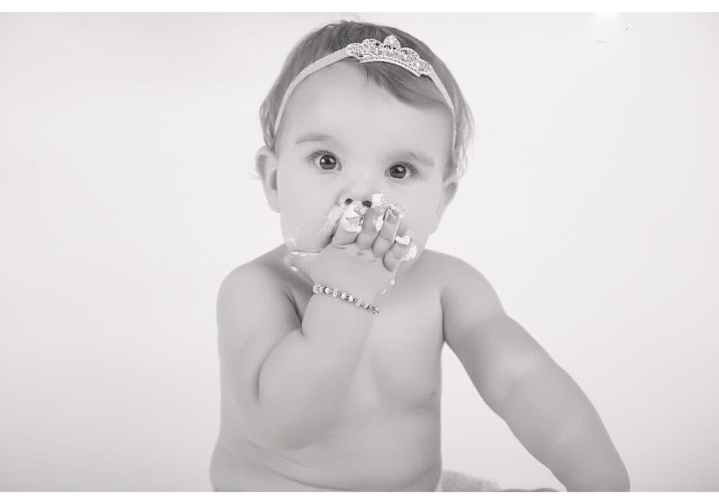 A baby wearing a headband and bracelet sits with a hand covered in cake or frosting, bringing it to their mouth. The photo is in black and white, and the baby looks directly at the camera with wide eyes. - Jodi O Photography