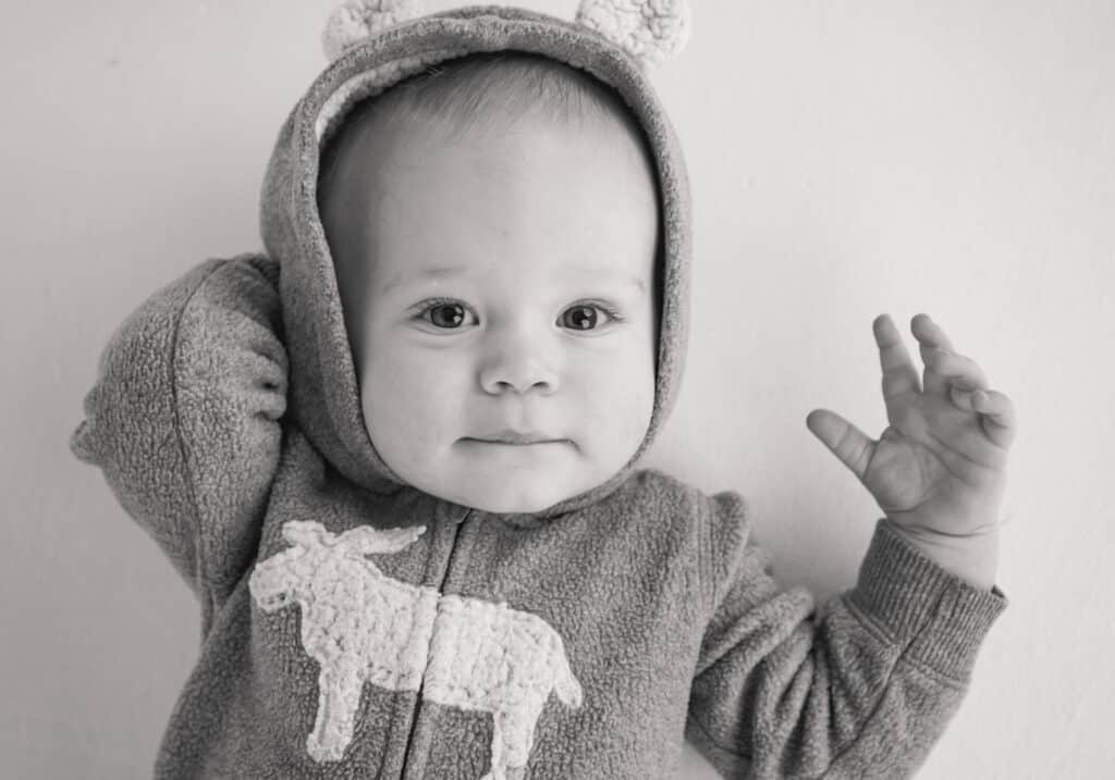 A baby wearing a hooded, fleece jacket with bear ears and a donkey design raises one hand and looks at the camera with a gentle expression. The photo is in black and white. - Jodi O Photography