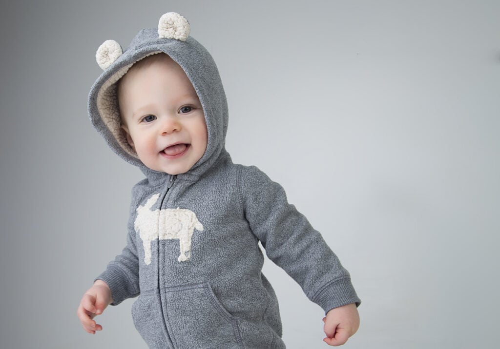 A smiling baby in a gray hooded jacket with bear ears and a white animal design stands against a light gray background. - Jodi O Photography