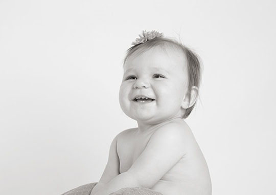 A black-and-white photo of a smiling baby with light hair, wearing a flower headband, seated and looking slightly to the side against a plain background. - Jodi O Photography