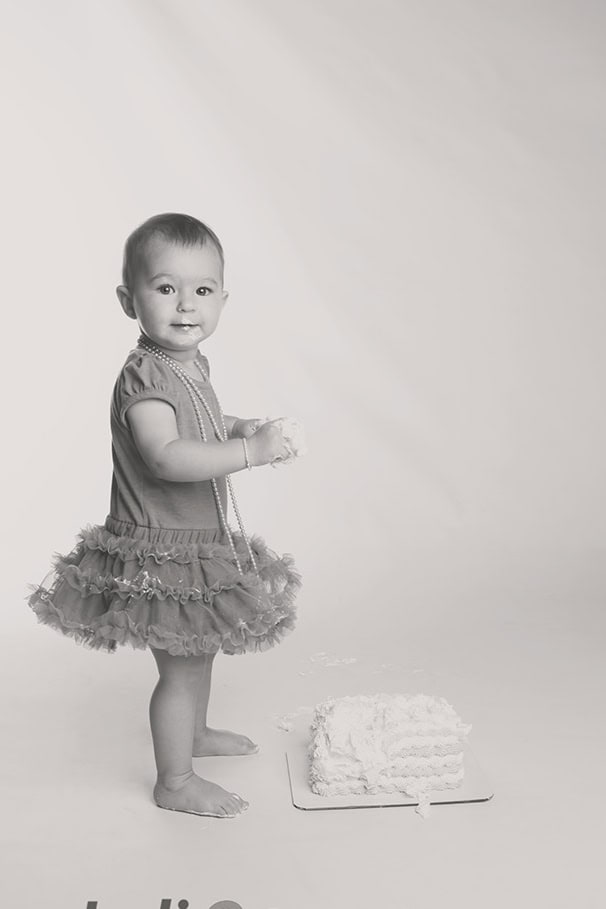 A smiling toddler in a frilly tutu and necklace stands barefoot beside a partially smashed cake, holding cake in her hands, against a plain studio background. - Jodi O Photography