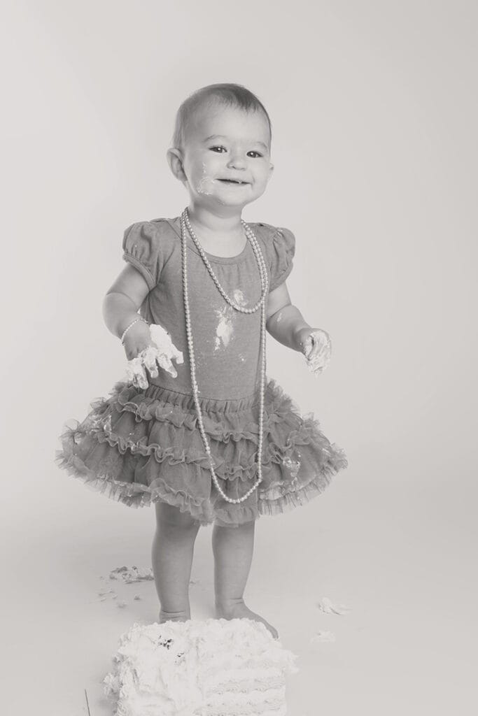 A smiling toddler in a ruffled dress and pearl necklace stands, with frosting on her hands and face, next to a smashed cake on the floor. The photo is in black and white. - Jodi O Photography
