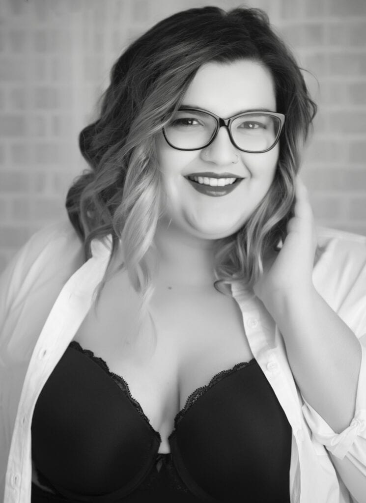 A smiling woman with wavy hair and glasses wears a white open shirt over a black bra, posing with one hand near her face. The photo is in black and white, with a blurred, tiled background. - Jodi O Photography