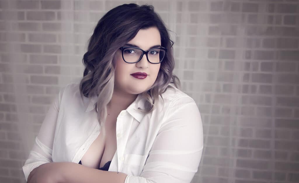A woman with wavy brown hair and glasses poses confidently in a white button-up shirt with a brick wall background. She has a calm expression and wears dark lipstick. - Jodi O Photography