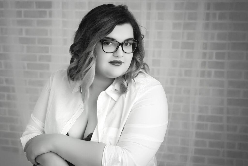 Black and white photo of a woman with wavy hair and glasses, wearing a white button-up shirt over a dark top, seated with her arms crossed, posing in front of a brick-patterned background. - Jodi O Photography