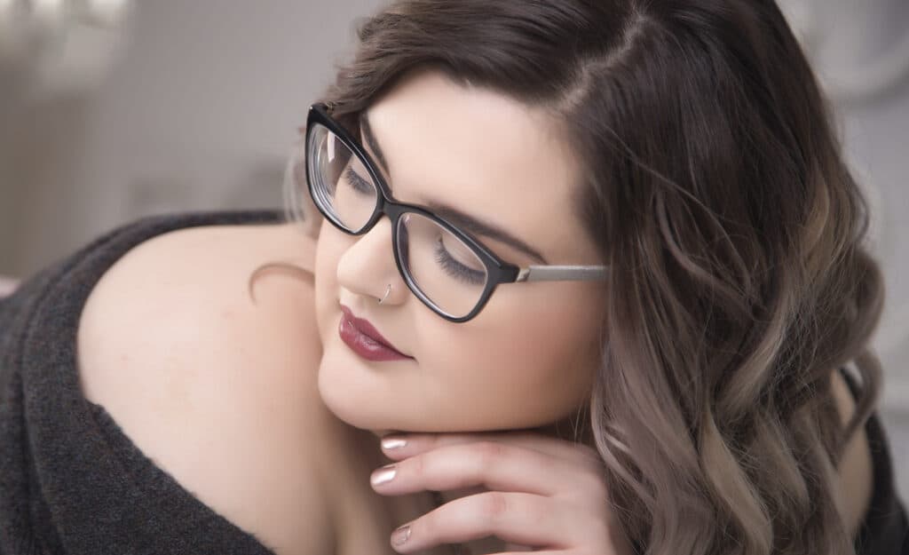 A woman with wavy brown hair, wearing glasses and a nose ring, rests her chin on her hand. She has soft makeup, dark lipstick, and is dressed in an off-shoulder top, looking down with a calm expression. - Jodi O Photography