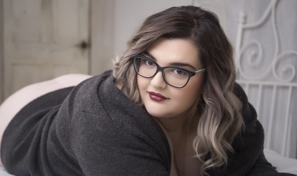 A woman with wavy, shoulder-length hair and glasses is lying on her stomach on a bed, wearing a dark sweater and looking directly at the camera with a slight smile. - Jodi O Photography