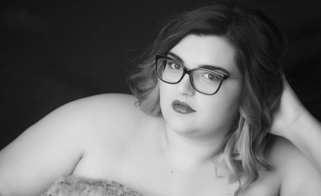 Black and white portrait of a person with shoulder-length hair, wearing glasses, looking at the camera with a calm expression. They are resting their head on one hand and wearing a strapless top. - Jodi O Photography