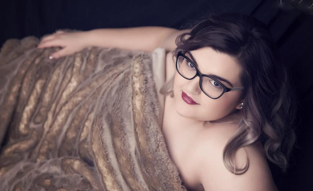 A woman with wavy brown hair and glasses looks up at the camera while wrapped in a soft, brown faux fur blanket against a dark background. - Jodi O Photography