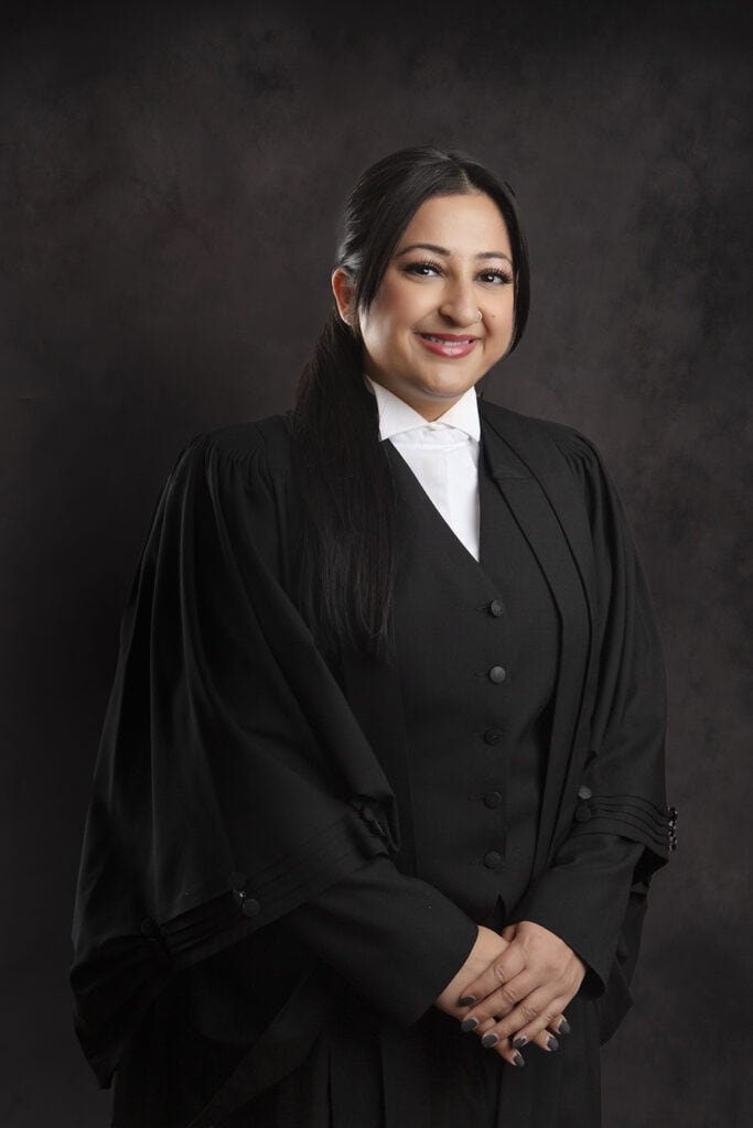 A woman in formal black legal attire stands against a dark background, smiling with her hands clasped in front of her. - Jodi O Photography