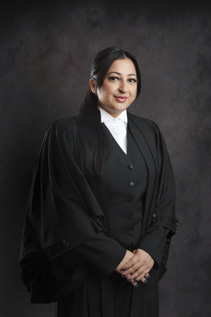 A woman with long dark hair, wearing a black legal robe and white collared shirt, stands against a dark, textured background, smiling with her hands clasped in front of her. - Jodi O Photography