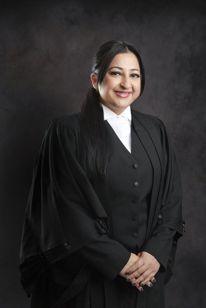 A woman in formal black legal attire stands against a dark backdrop, smiling with her hands clasped in front of her. She has long dark hair pulled back and is wearing a white shirt and black robes, suggesting she is a lawyer or judge. - Jodi O Photography