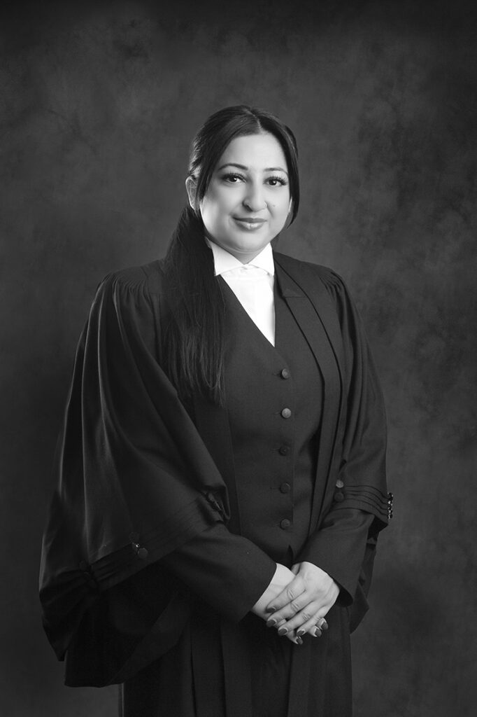 Black and white portrait of a woman wearing formal legal attire, including a robe and white collar, standing against a dark, textured background with her hands clasped in front of her and a gentle smile. - Jodi O Photography