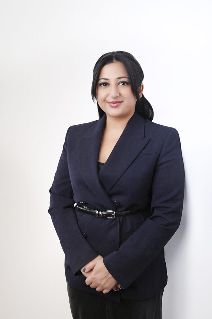 A woman with long dark hair tied back, wearing a navy blazer and black top, stands against a plain white background, smiling slightly with her hands clasped in front of her. - Jodi O Photography