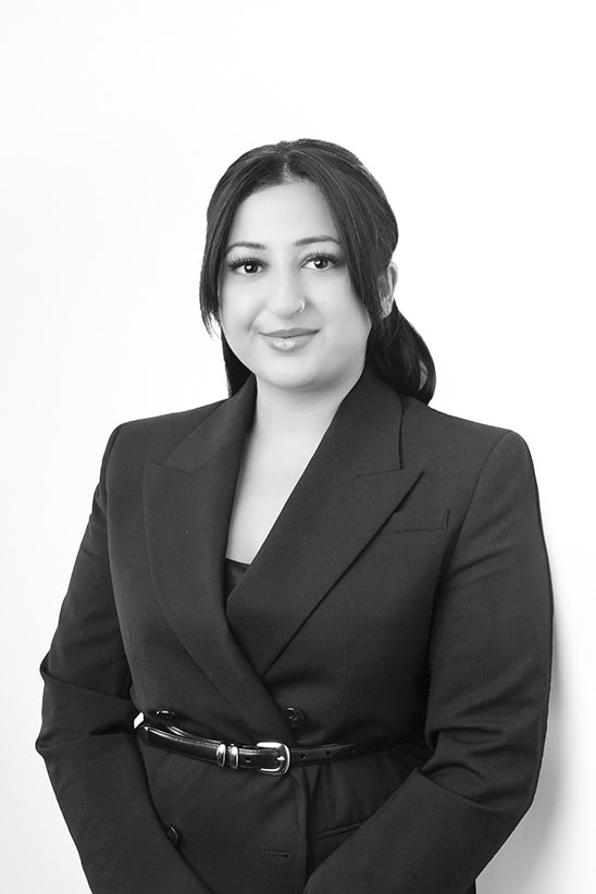 A woman with long dark hair, styled back, wearing a dark blazer with a belt, stands against a plain white background, smiling gently at the camera. The image is in black and white. - Jodi O Photography