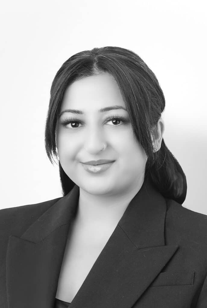 Black and white portrait of a woman with dark hair in a low ponytail, wearing a blazer, smiling slightly, and facing the camera against a plain background. - Jodi O Photography