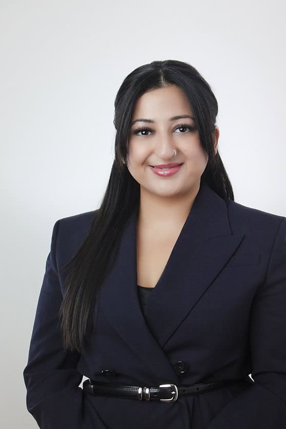 A woman with long, dark hair styled straight, wearing a dark blazer and black top, stands against a plain light background, smiling at the camera. - Jodi O Photography