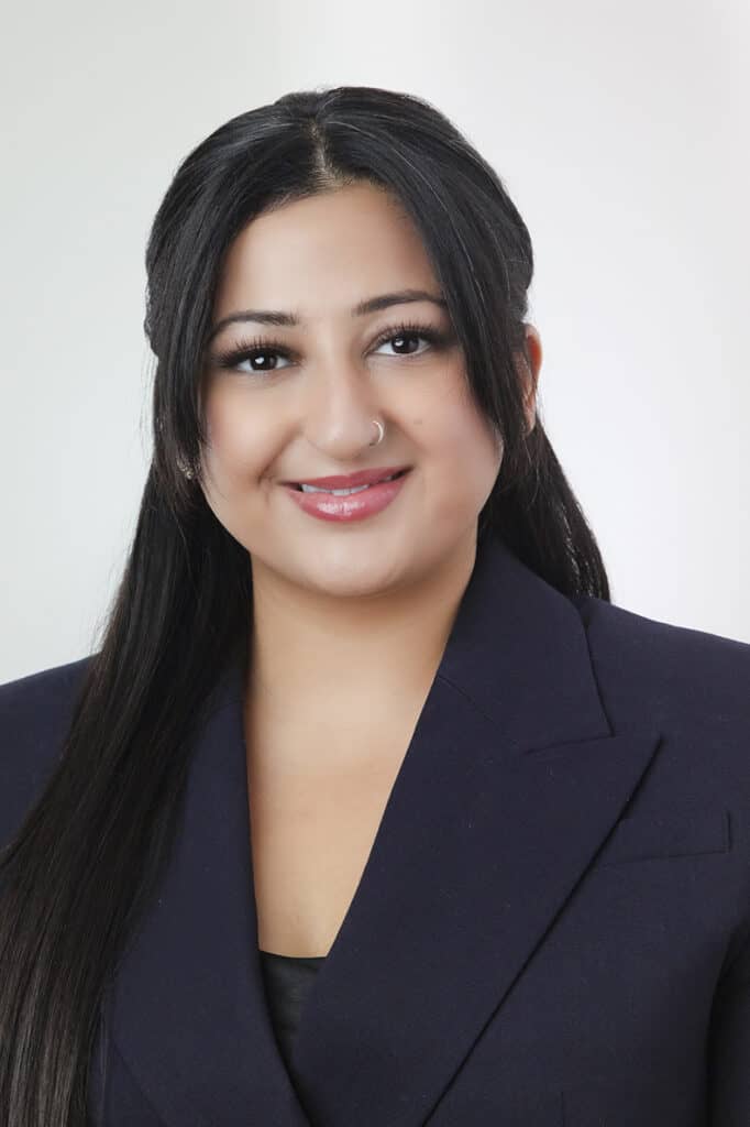 A woman with long, straight dark hair, wearing a dark blazer and a black top, smiles at the camera against a plain light background. - Jodi O Photography